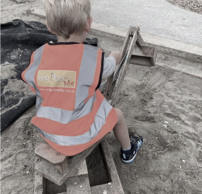 Child sitting down on a wooden digger wearing a Pro-Tect Me high-vis orange jacket